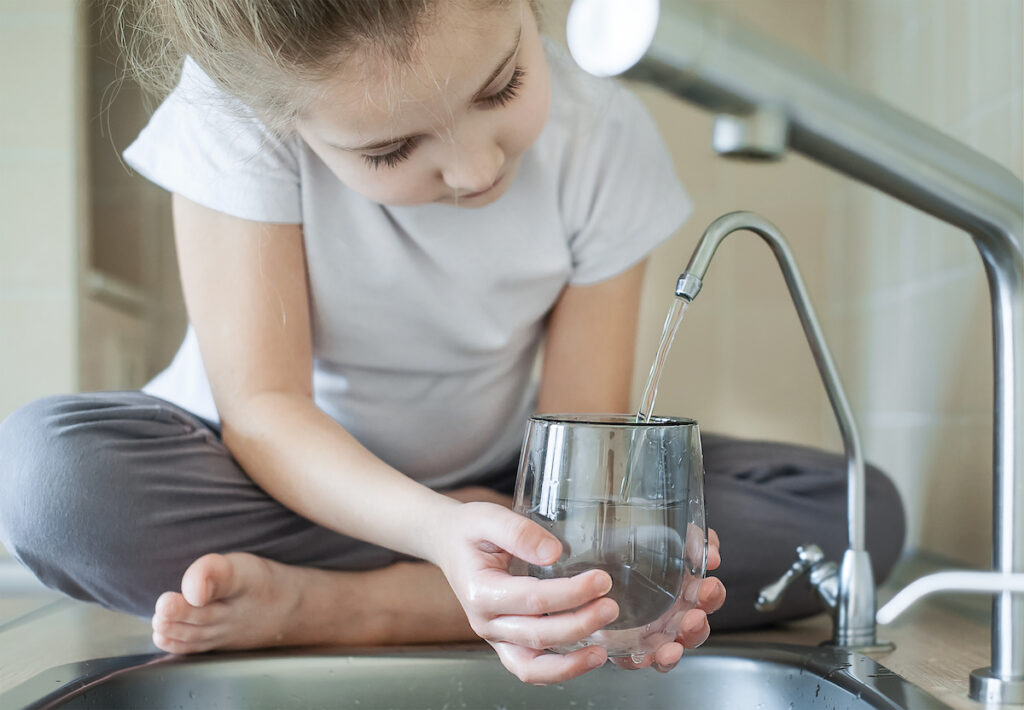 Little girl open a water tap with her hand holding a transparent glass. Kitchen faucet. Filling cup beverage. Pouring fresh drink. Hydration. Healthcare. Healthy lifestyle. Reverse osmosis system