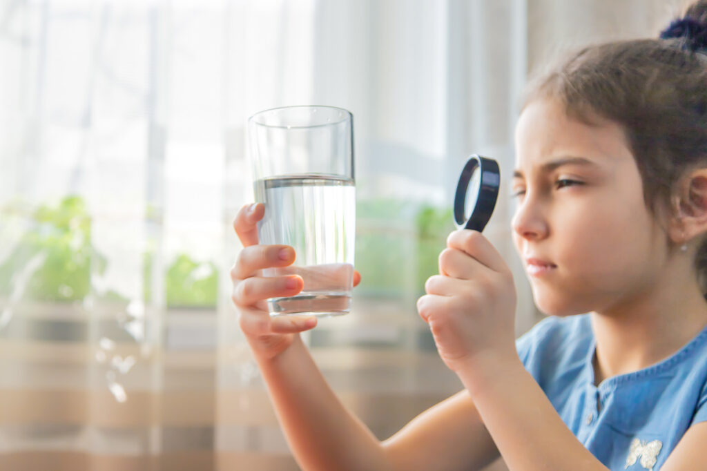 A young girl holding a magnifying glass up to a glass of water to check for water filtration.