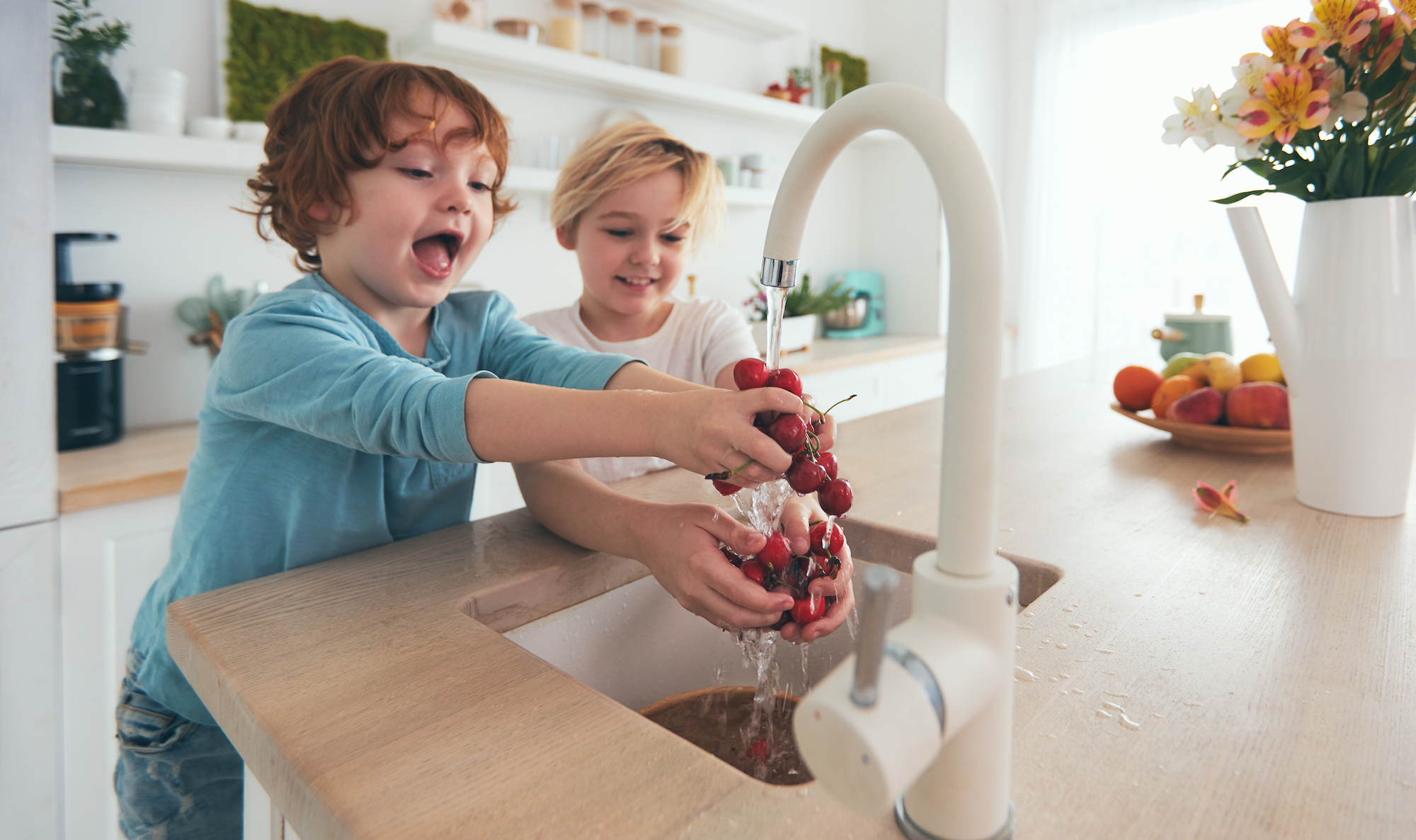 Two young children washing handfuls of cherries with tap water from the kitchen sink